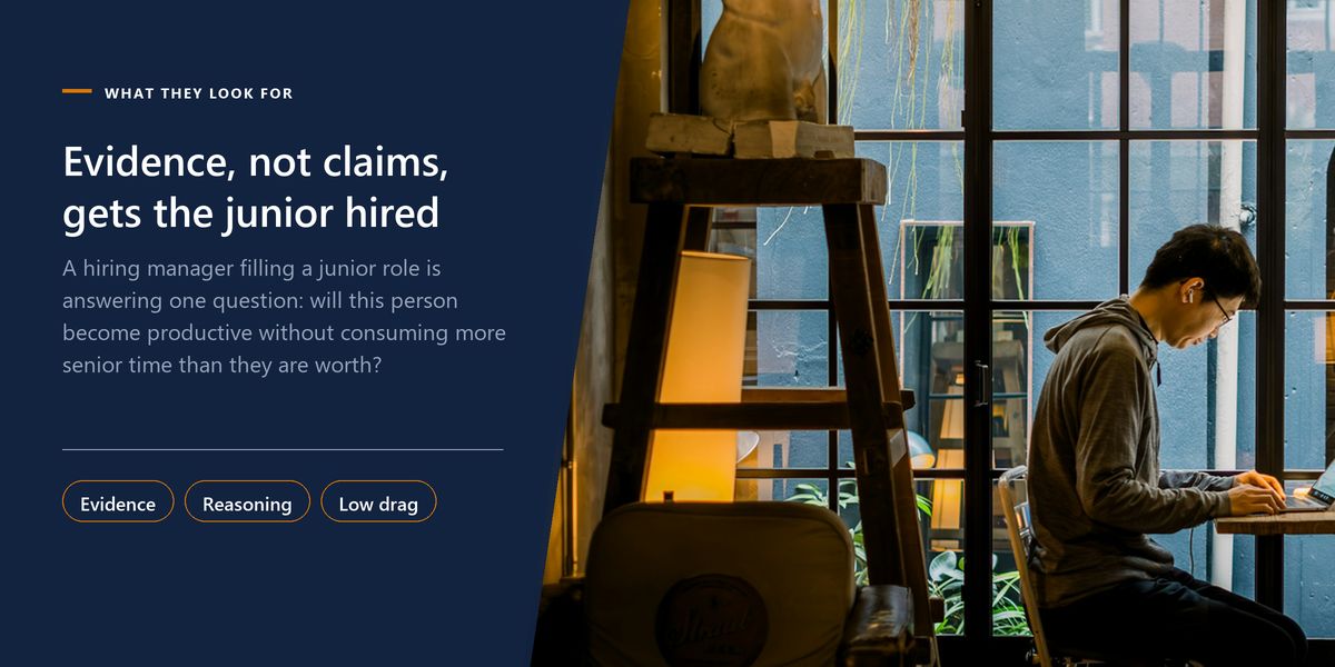 A man works on a laptop in a cafe window at dusk, beside a banner stating that evidence, not claims, gets the junior hired.